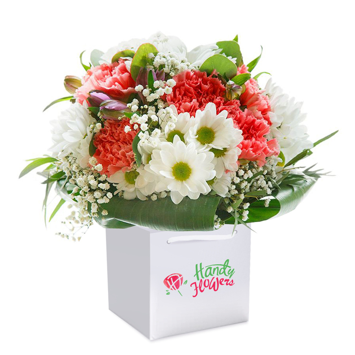 Set against a pristine white background, this exuberant bouquet exudes freshness and vitality in the quintessential style of Bloomsbury. The focal point is a generous gathering of peach and coral carnations, their ruffled petals layered in abundance, creating a sumptuous sense of nurture and warmth. Between them, crisp white daisies sparkle, edged with subtle green and bearing sunny yellow-green hearts reminiscent of the springtime moods in Bloomsbury's historic gardens. Sprigs of fine, white baby's breath mingle with the main blooms, their airy form invoking a feeling of lightness and approachability often found in local community spaces. Encircling the arrangement, glossy, deep green leaves unfold with gentle intent, sculpting a harmonious silhouette and reinforcing the bouquet's rounded shape. On one side emerges a shy group of purple-tinged buds, offering an undercurrent of excitement and anticipation akin to finding a hidden alcove in the British Museum. The sturdy white carrier bag, finished with soft ribbon handles, promises both convenience and contemporary style, devoid of overt branding so the flowers remain the star. Every detail-colours, forms, composition-is expertly chosen, revealing the care and tradition of Bloomsbury Florist. Ideal as a birthday surprise, an anniversary keepsake, or simply to uplift the everyday, this arrangement celebrates both handcrafted artistry and the cultured charm of London's literary quarter.